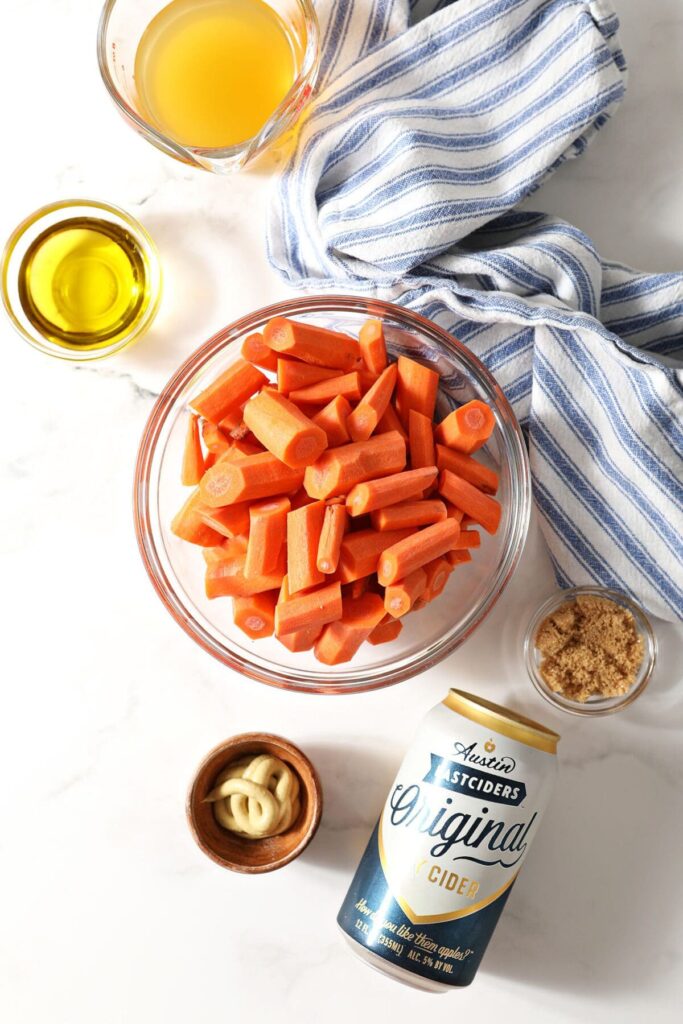 Carrots in a bowl and other ingredients to make cider glazed carrots on marble with a blue striped towel