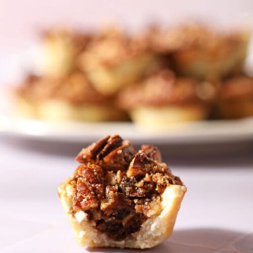 a bitten-into pecan tassies sits on a purple tiled surface in front of a white plate of more