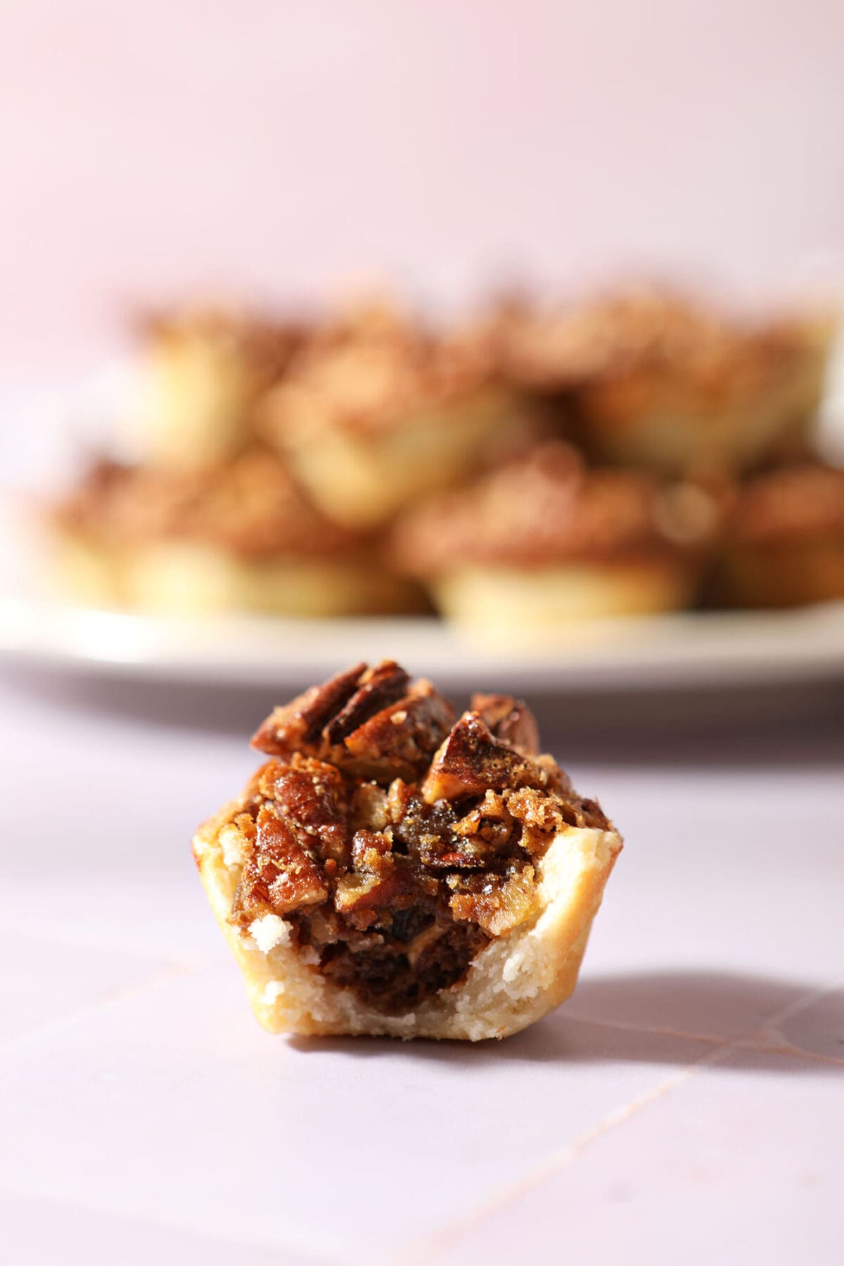 a bitten-into pecan tassies sits on a purple tiled surface in front of a white plate of more