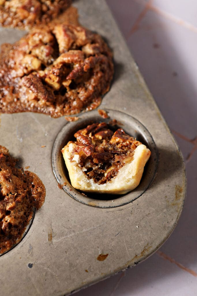 a bitten-into pecan tassie sits in a mini muffin tin with more tassies