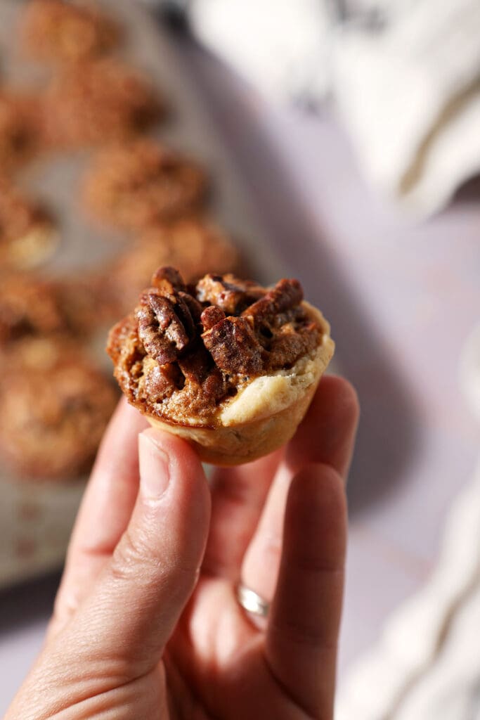 a hand holds a pecan tassie above a mini muffin tin of more tassies on a purple surface