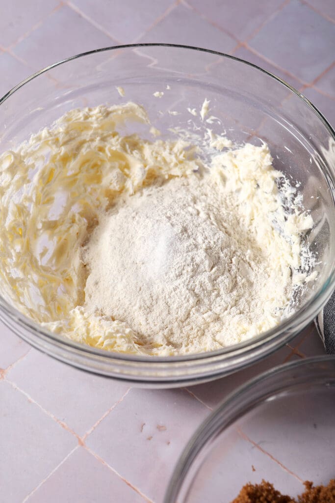 flour and salt sit on top of creamed cream cheese and butter in a bowl on a purple surface