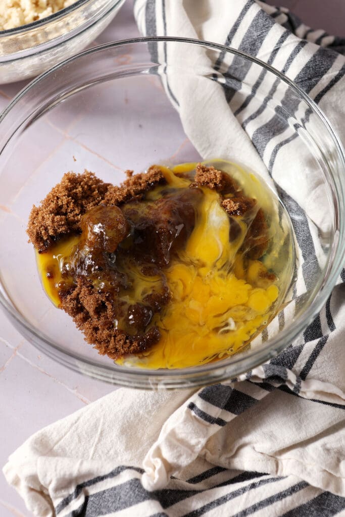 eggs and brown sugar in a bowl before mixing on a light purple surface next to a white and black striped linen