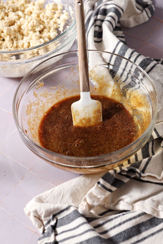 mixed egg and brown sugar in a bowl on a light purple surface next to a white and black linen and other ingredients