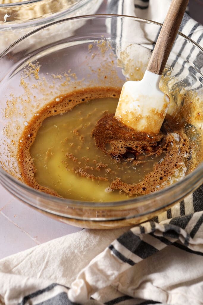 butter sits on top of other liquid ingredients for a pecan pie cookie recipe in a bowl on a light purple surface