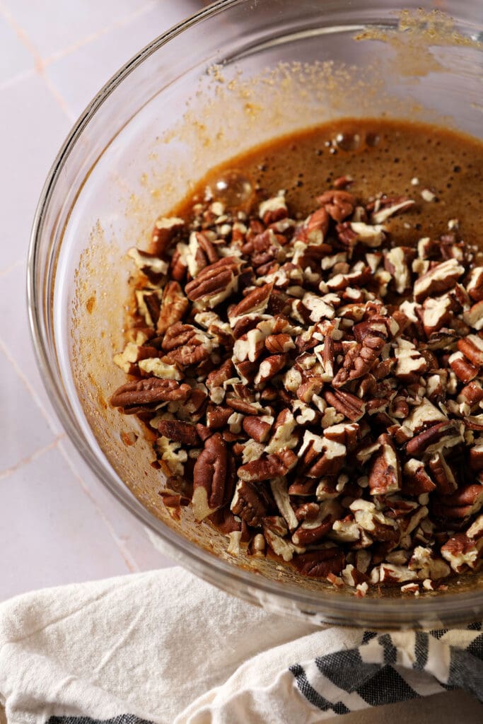 chopped pecans sit on top of liquid ingredients for pecan tassie cookies in a bowl on a light purple surface