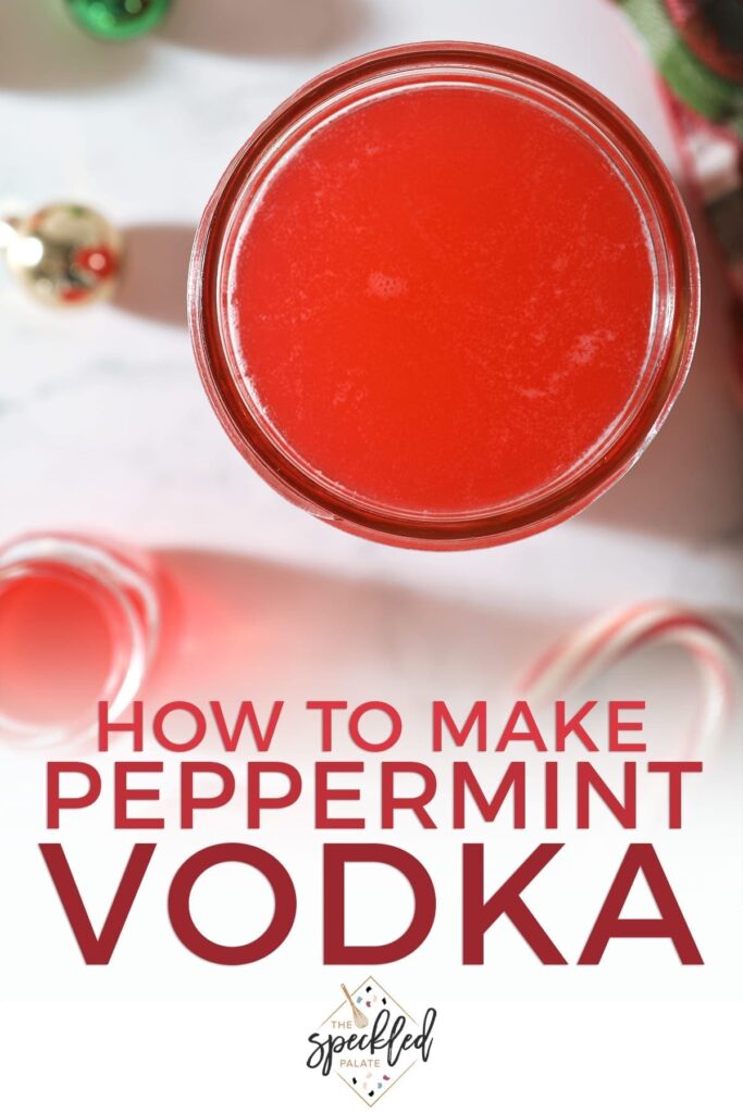 Close up of a mason jar of peppermint vodka next to a two shot glasses with glass ornaments, a Christmas-colored plaid towel and a candy cane on marble with the text 'how to make peppermint vodka'