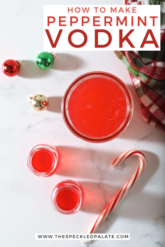 Two shot glasses of peppermint vodka sit next to a mason jar full of the candy cane-infused vodka with glass ornaments, a Christmas-colored plaid towel and a candy cane on marble with the text 'how to make peppermint vodka'