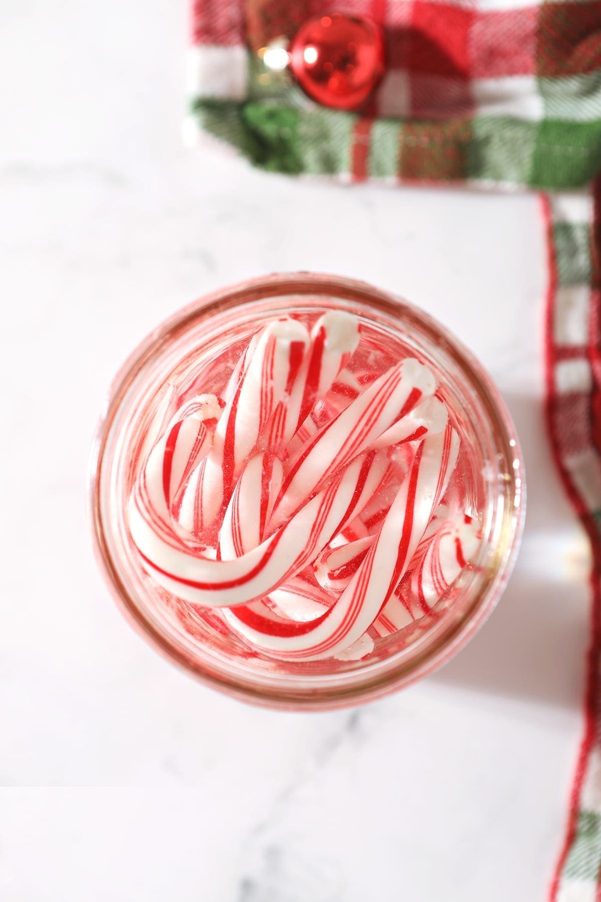 Several candy canes in a mason jar full of vodka next to a green and red plaid kitchen towel on a marble surface