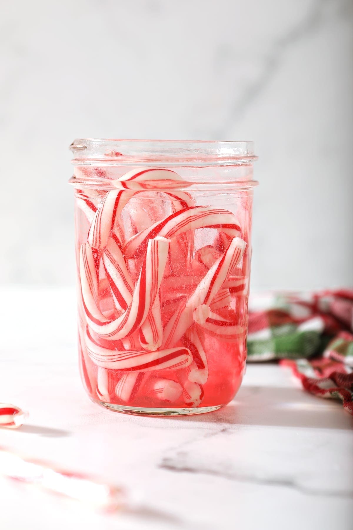 Several candy canes melt into a mason jar full of vodka next to a green and red plaid kitchen towel on a marble surface