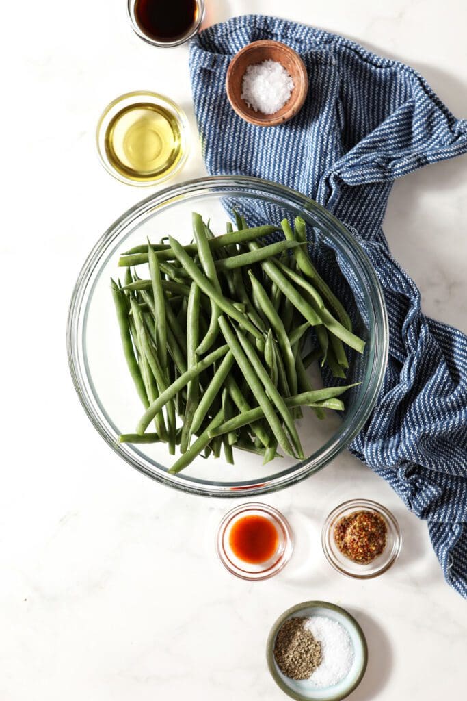 Green beans in a bowl surrounded by dressing ingredients on marble with a blue linen