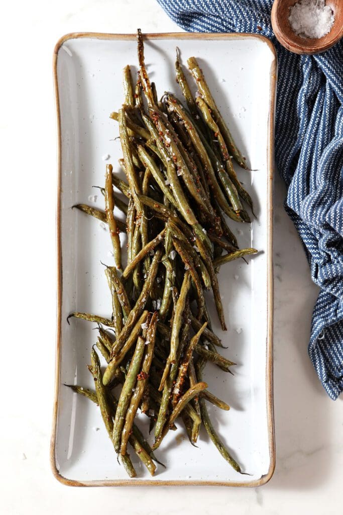 Roasted green beans on a white platter next to a blue linen on marble