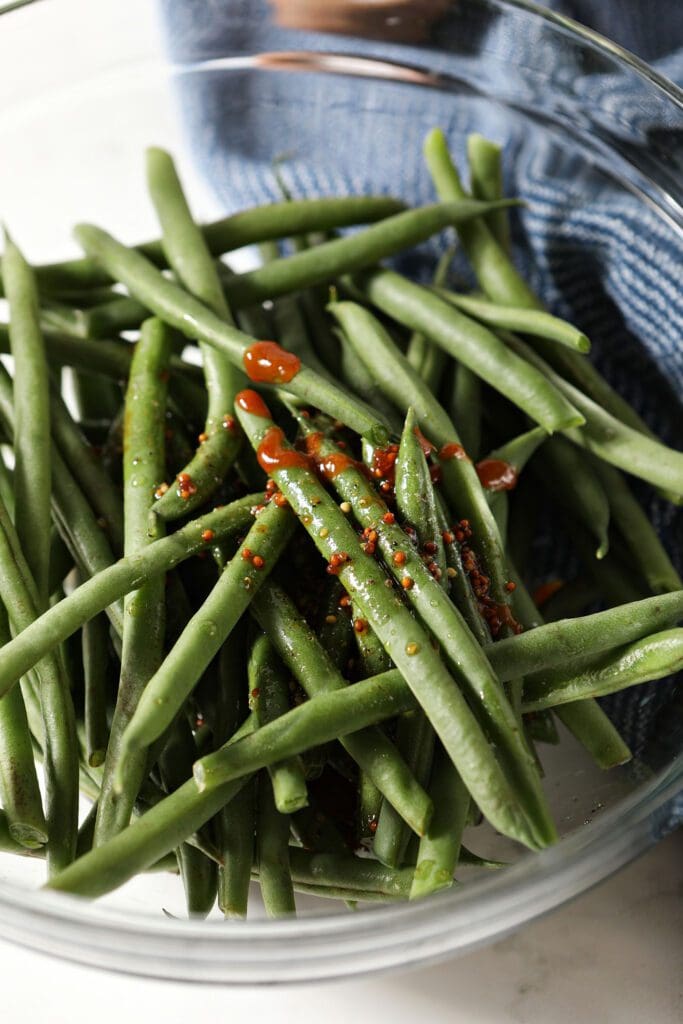 A bowl of green beans topped with a dressing next to a blue linen