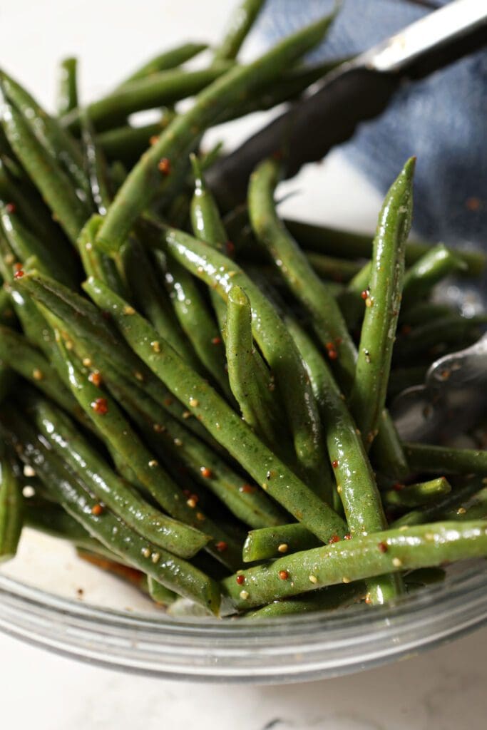 Green beans are tossed in a dressing with tongs in a glass bowl