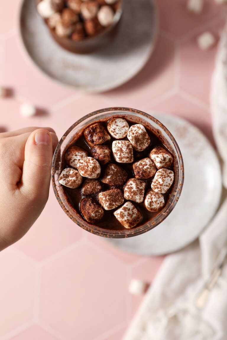 A child holds a mug of Dairy Free Hot Chocolate topped with marshmallows and cocoa powder over a plate and a pink tiled surface