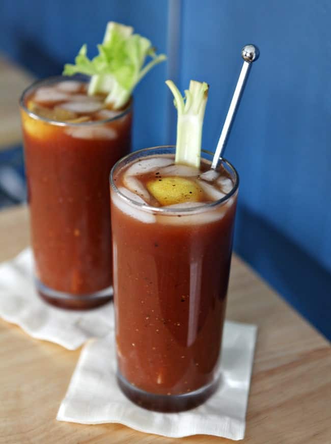 Two tall glasses of Bloody Marys are shown on a wooden background next to a blue wall