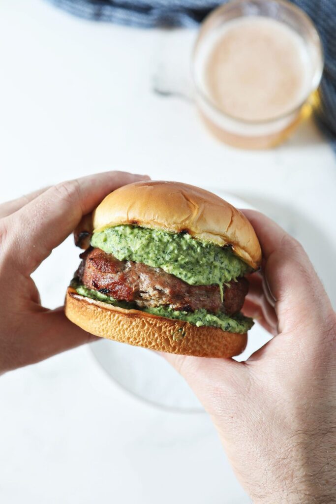 A man holds a burger topped with spinach-artichoke dip above a plate