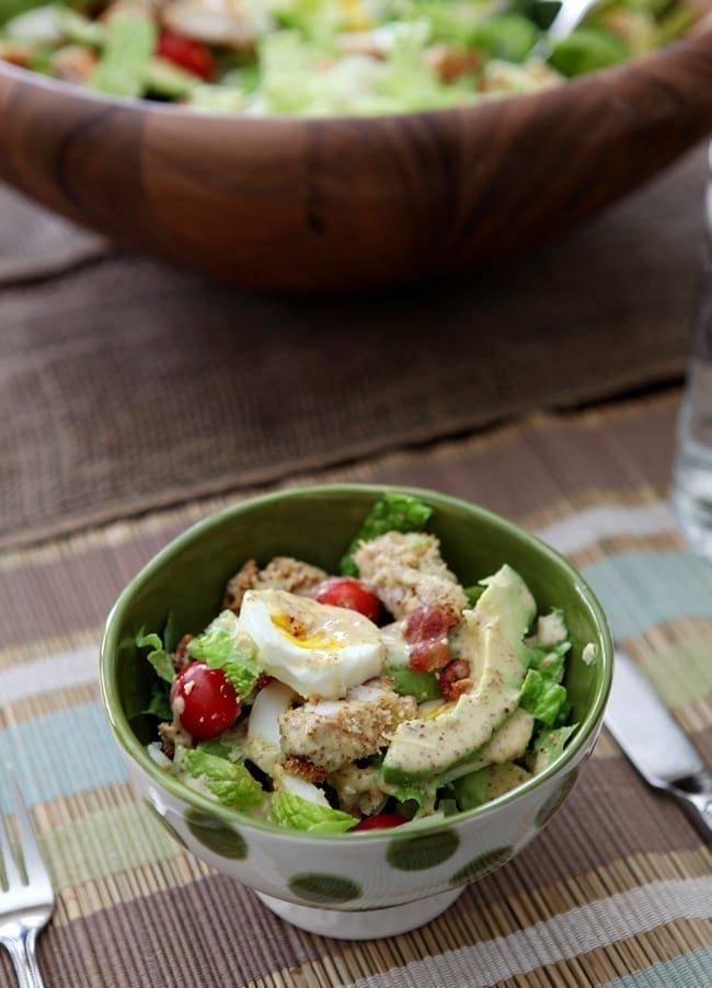 A bowl of Avocado and Panko-Breaded Chicken Cobb Salad sits on a table with the serving bowl of the salad in the background.