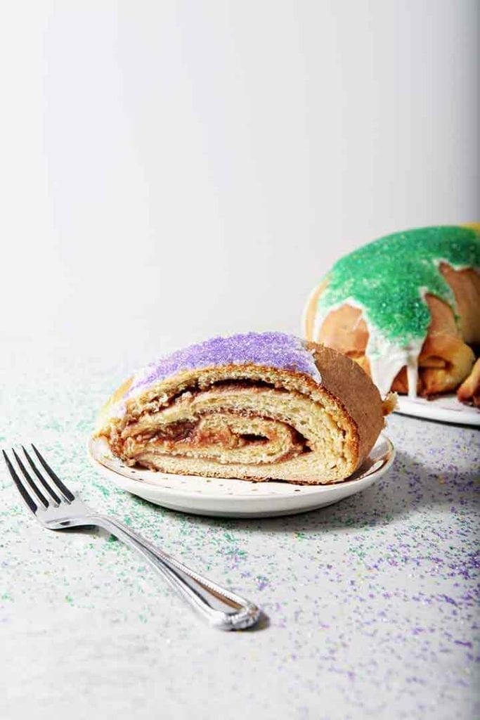 A slice of Cream Cheese and Raspberry King Cake sits in front of the whole King Cake