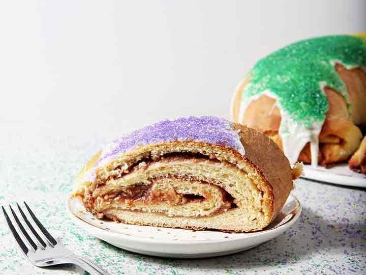 A slice of Cream Cheese and Raspberry King Cake sits in front of the whole King Cake