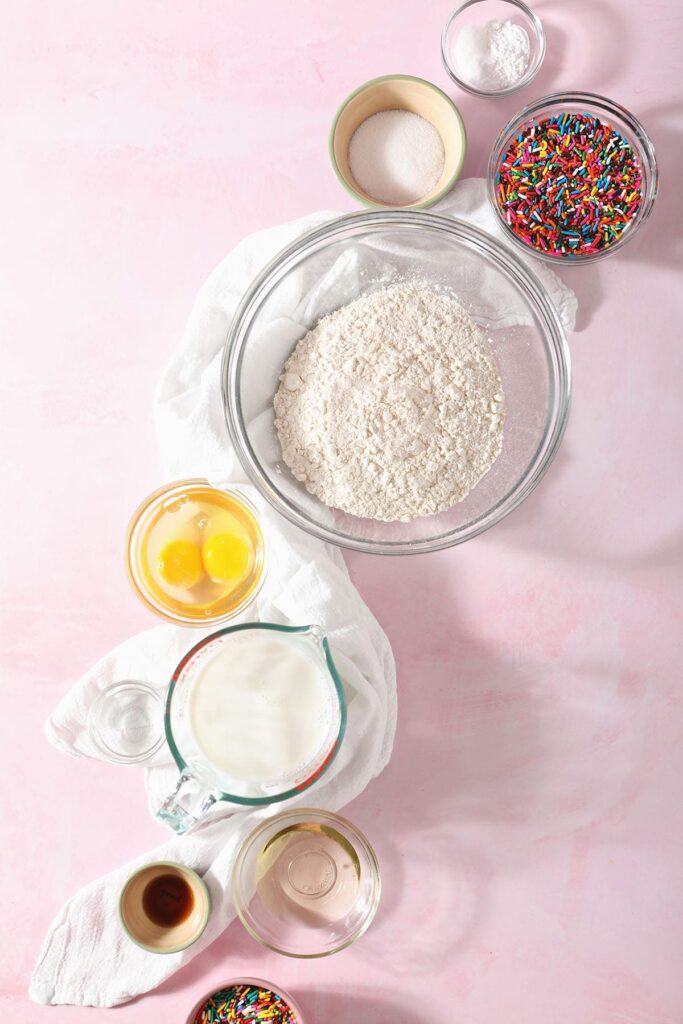 Ingredients for birthday cake pancakes in bowls on a pink countertop