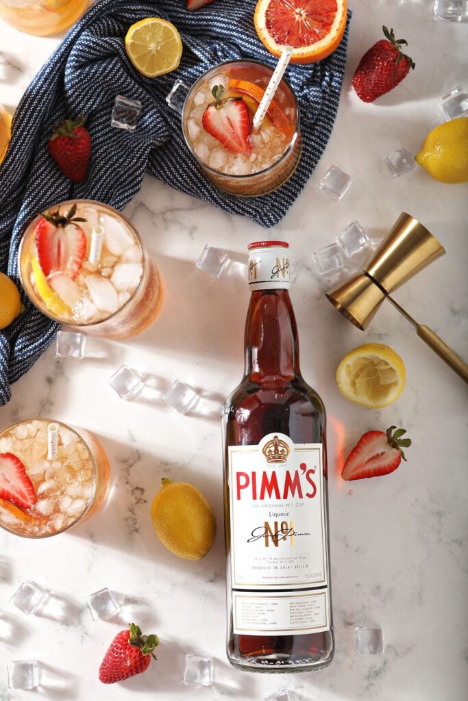 Five fruity drinks garnished with strawberry sit next to a bottle of Pimm's No. 1 on marble