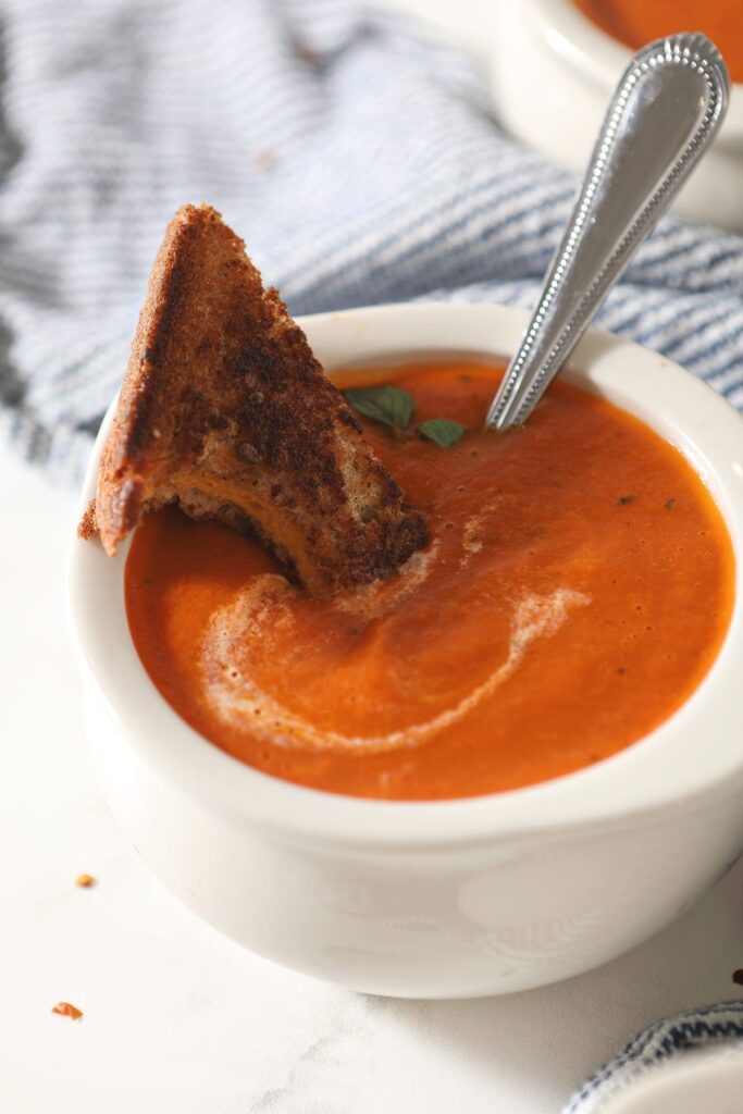 A half-bitten triangle of a grilled cheese sandwich sits on top of a white bowl holding creamy tomato soup