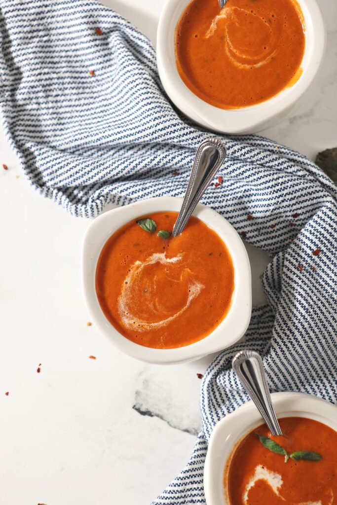 Three white bowls of tomato soup with spoons in them sit on a striped blue towel