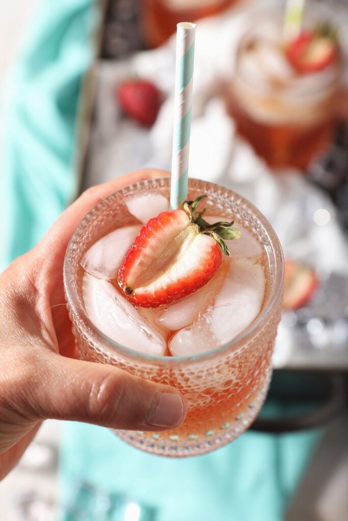 A person holds a glass of homemade strawberry tea garnished with a strawberry half