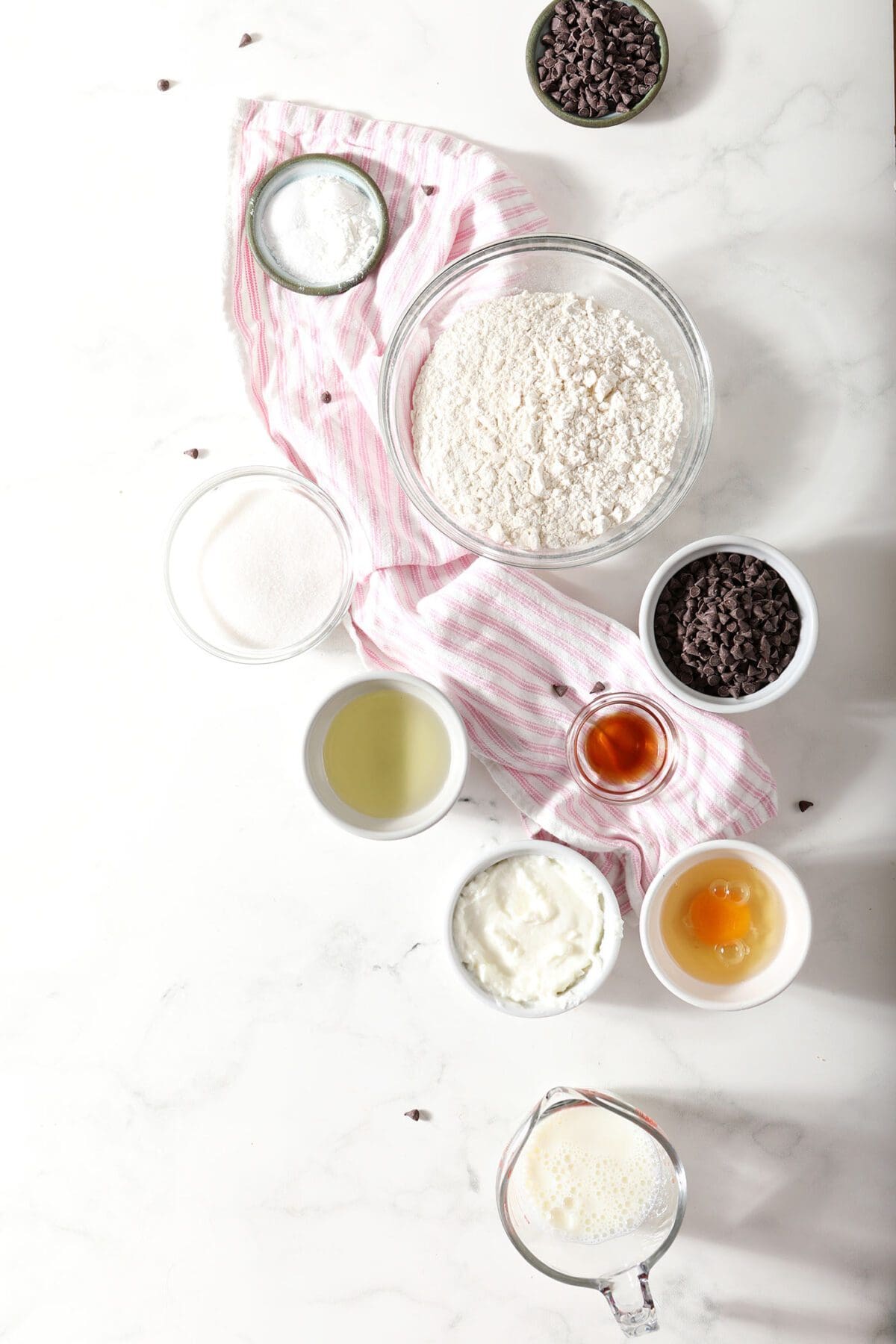 Ingredient to make muffins with chocolate chips in bowls on a marble countertop with a pink striped towel