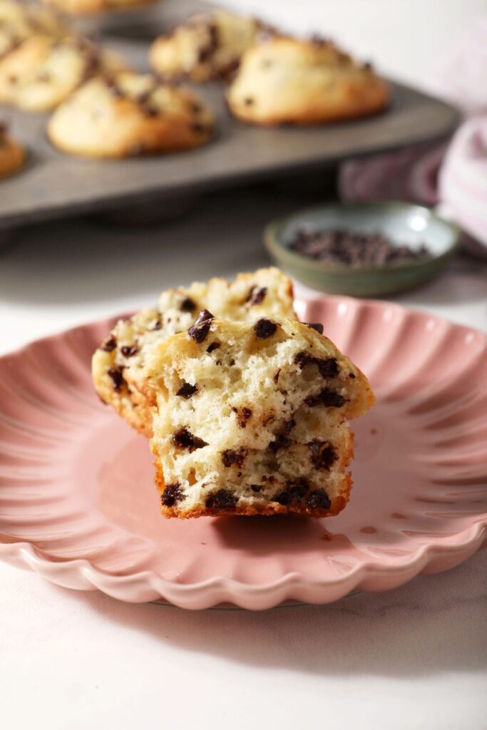 A halved chocolate chip muffin on a pink plate in front of a muffin tin