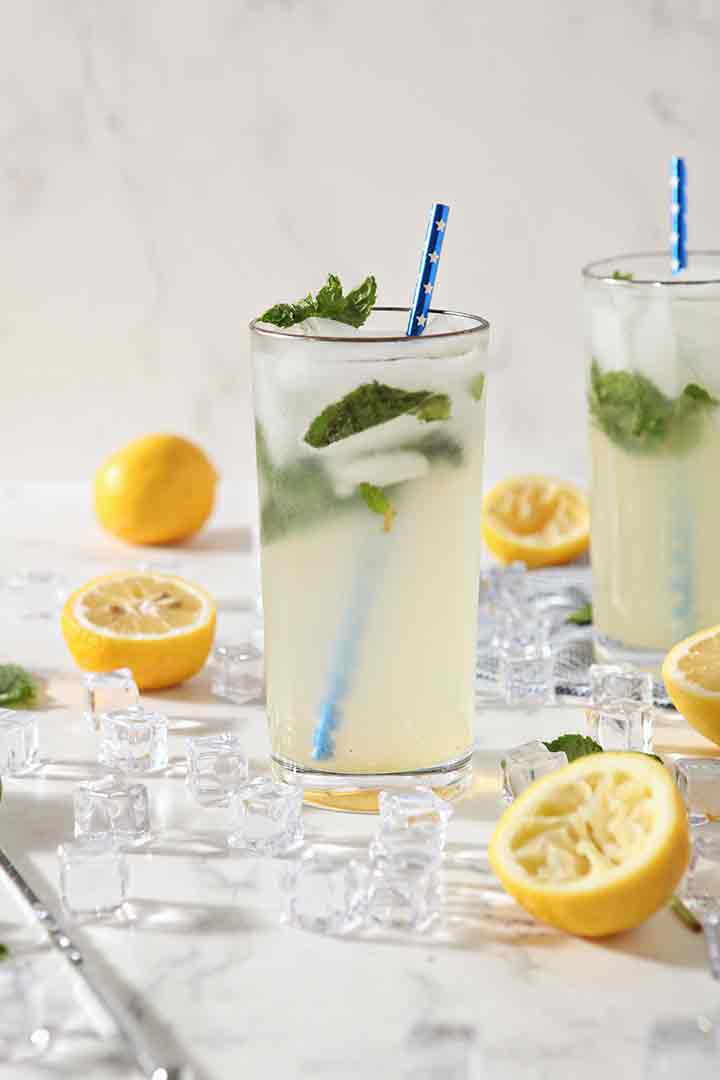 Two glasses of Mint Lemonade are ready for drinking, surrounded by ice, lemons and mint leaves