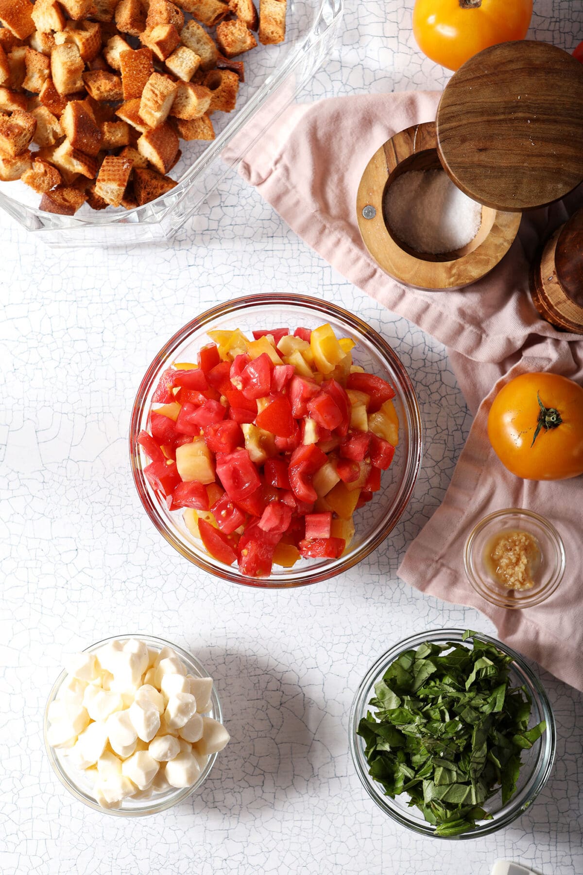 ingredients to make caprese-style panzanella in bowls on a white surface with a pink linen