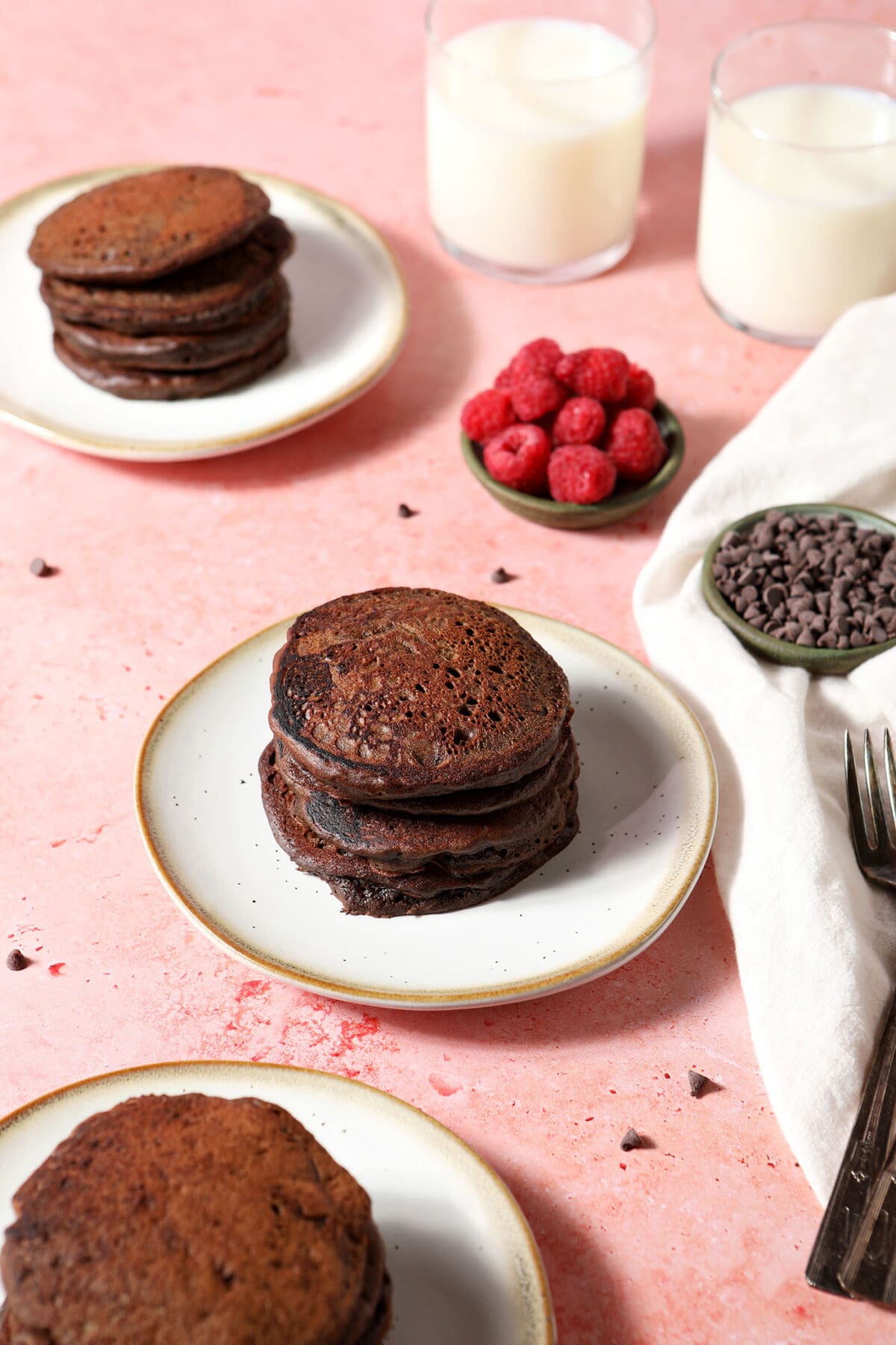 Three white plates with stacks of chocolate pancakes on a pink surface with bowls of raspberries and chocolate chips and cups of milk