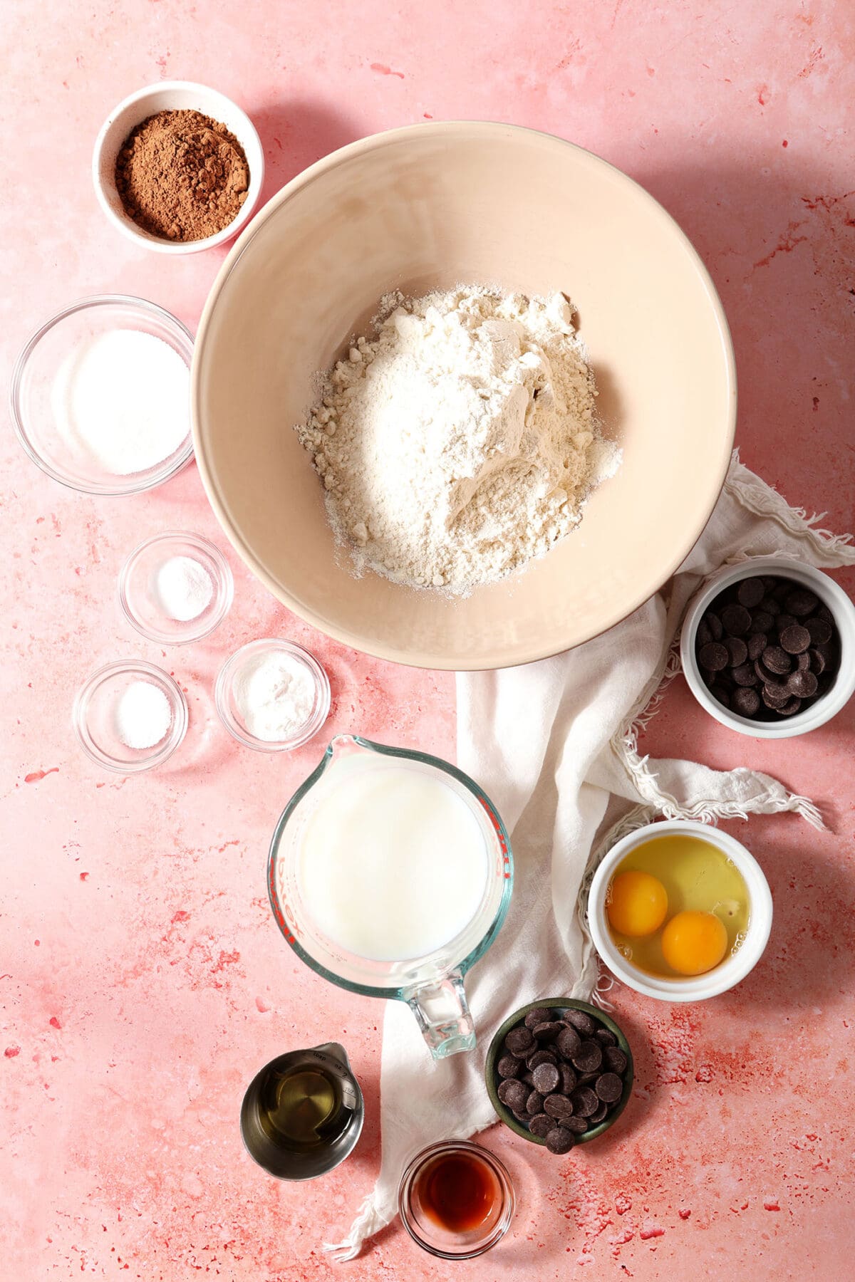 Ingredients to make chocolate pancakes in bowls on a pink tiled surface with a white linen