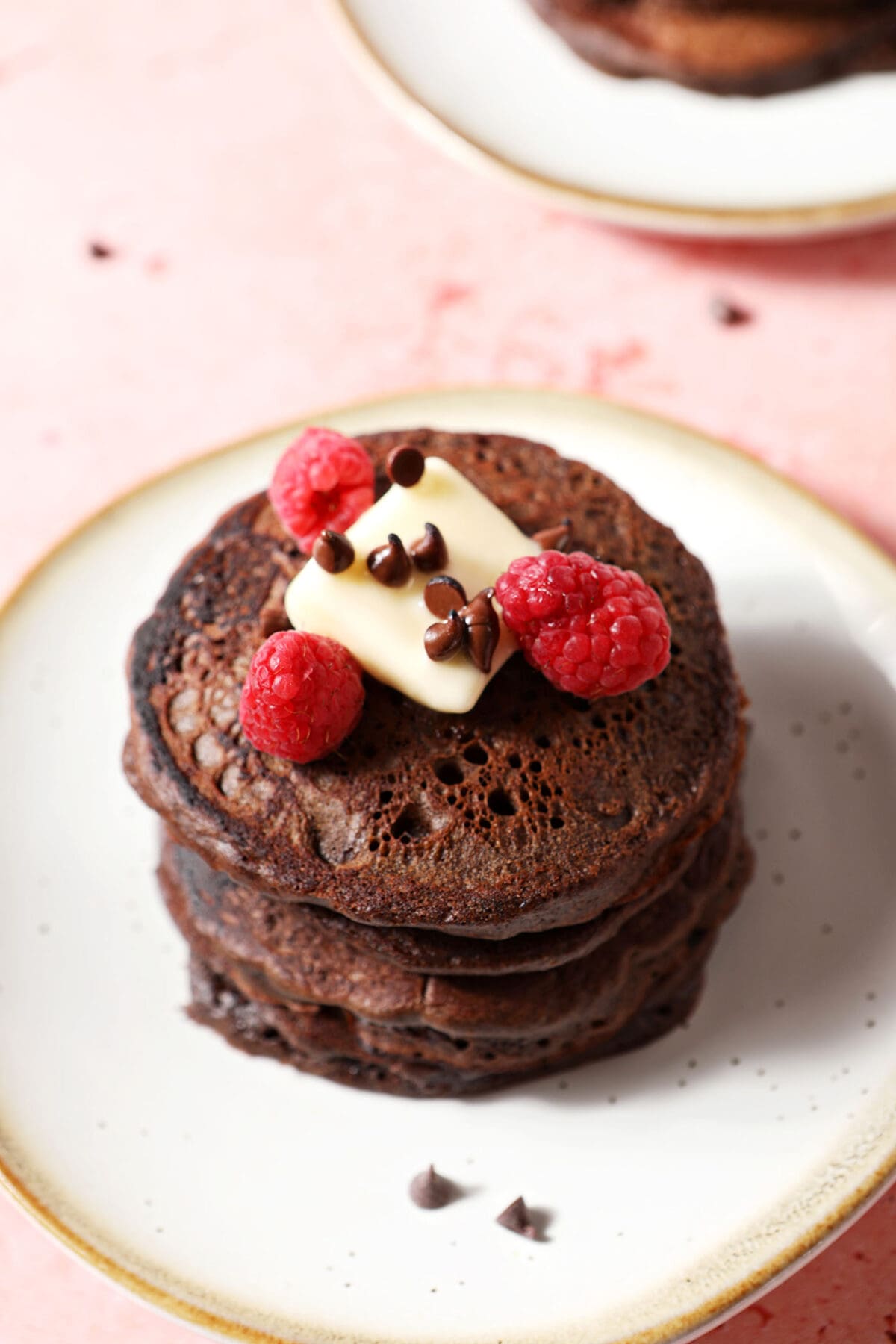 A stack of Chocolate Pancakes topped with a pat of butter, mini chocolate chips and raspberries on a white plate on a pink surface