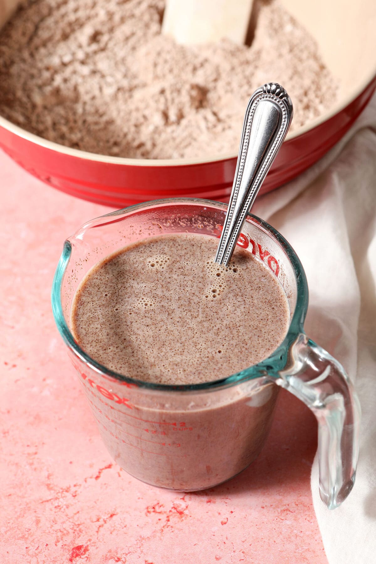 Chocolate milk and other liquid ingredients in a cup on a pink surface next to a bowl of dry ingredients to make pancakes