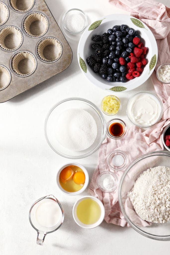 Bowls of ingredients to make muffins with mixed berries on a white surface with pink linens