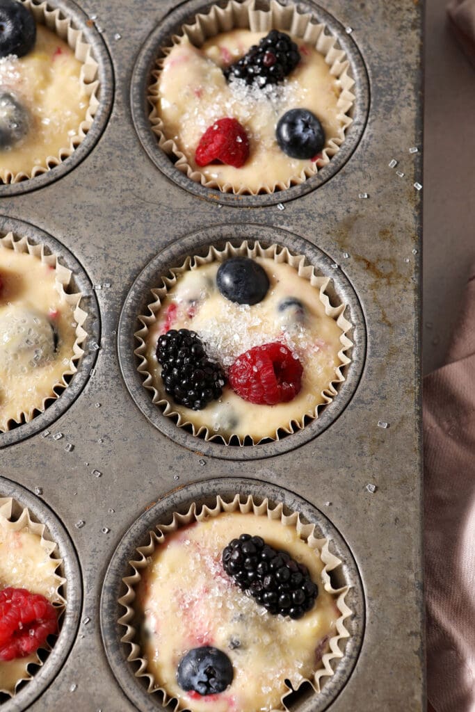 Muffin batter topped with raspberries, blueberries and blackberries and sparkling sugar in muffin tins before baking