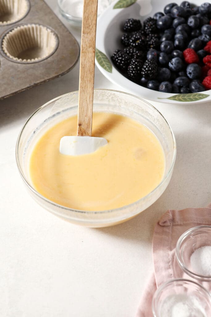 Wet ingredients for muffins in a bowl next to mixed berries in a bowl and dry ingredients