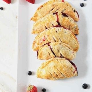 Strawberry and Blueberry Hand Pies sit on a white platter, surrounded by fresh blueberries and strawberries, before serving.