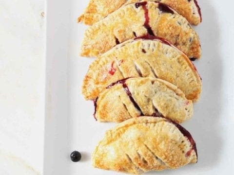 Strawberry and Blueberry Hand Pies sit on a white platter, surrounded by fresh blueberries and strawberries, before serving.