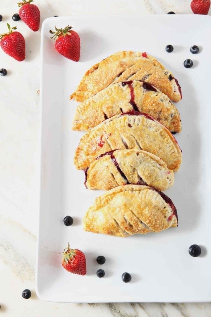 Strawberry and Blueberry Hand Pies sit on a white platter, surrounded by fresh blueberries and strawberries, before serving.