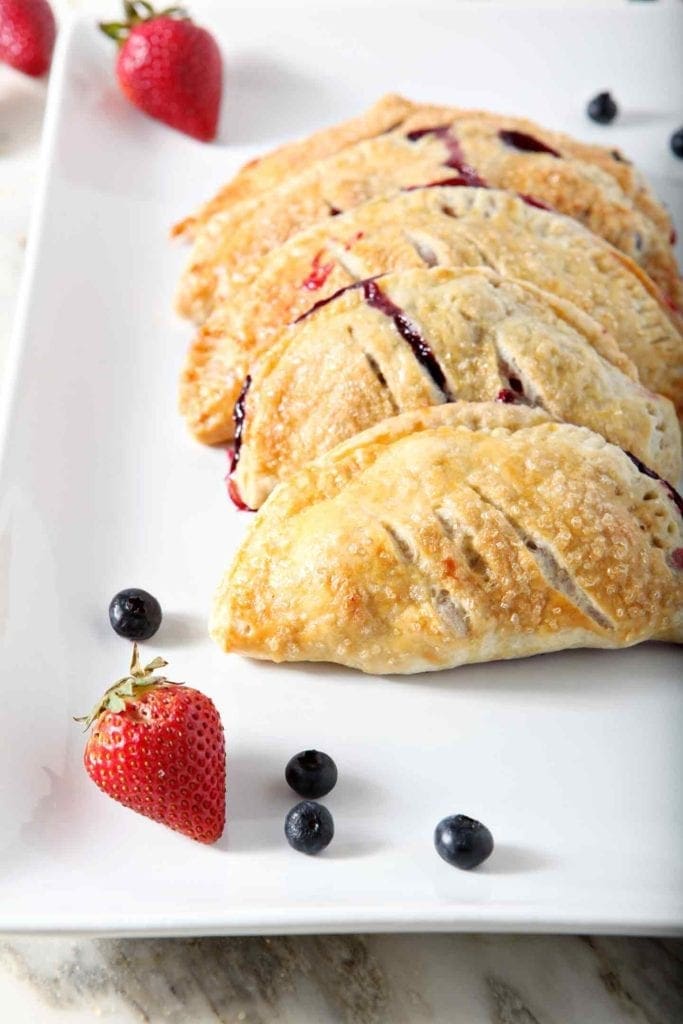 Several Hand Pies are stacked on a plate, surrounded by fresh strawberries and blueberries, before serving