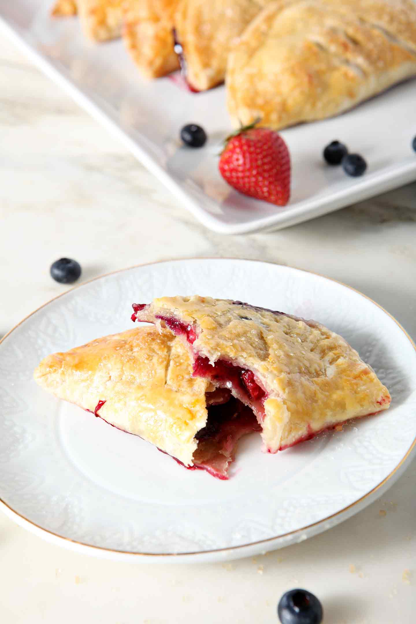 A Strawberry and Blueberry Hand Pie, broken in half, sits on a white dessert plate, with a platter of additional hand pies behind it.