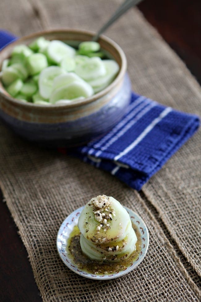 A small bowl holds a stack of cucumber slices topped with a feta vinaigrette on a tabletop with a bowl holding cucumbers behind it