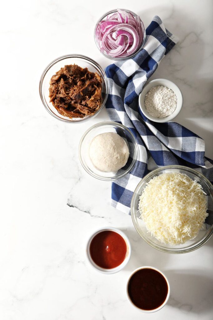 Ingredients to make homemade bbq pizza in bowls on marble with a blue buffalo check towel