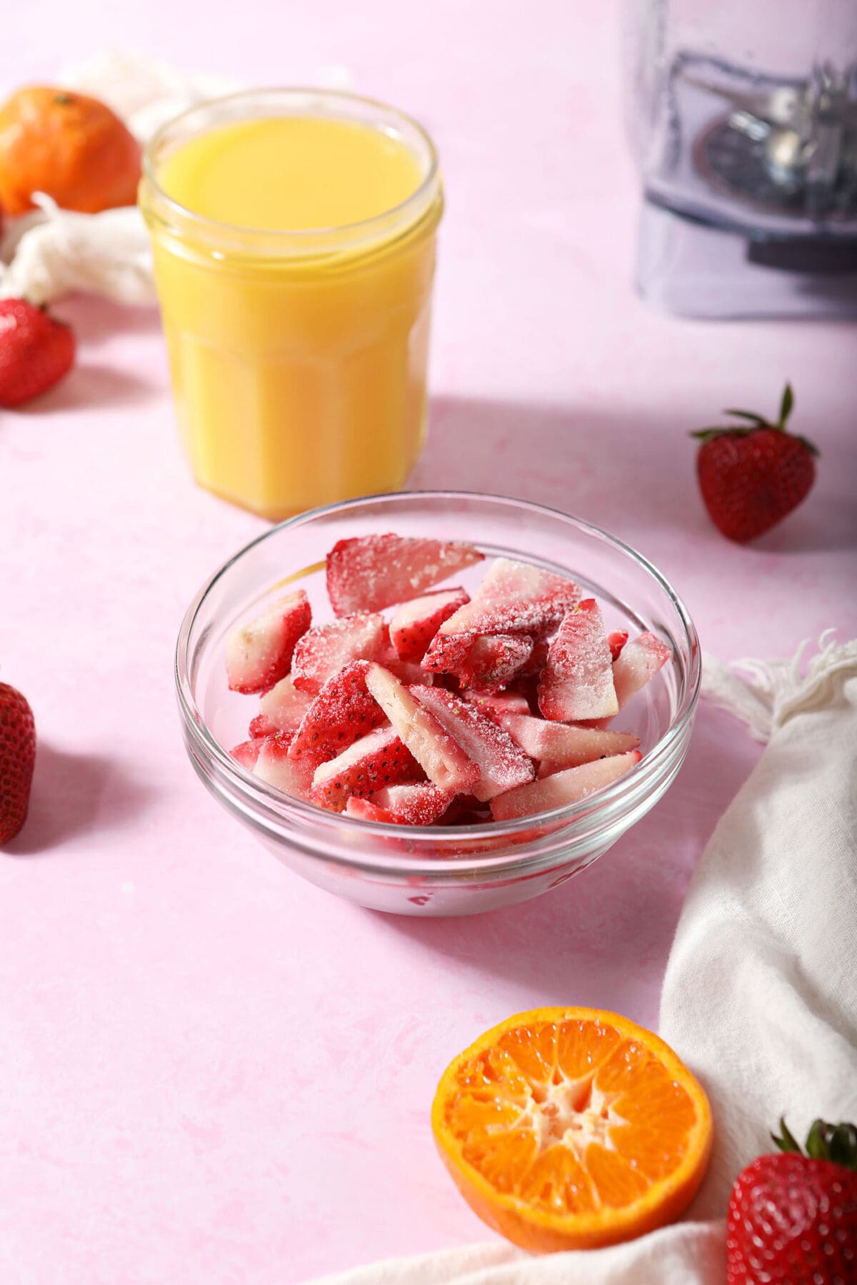 frozen strawberries in a bowl next to a glass of orange juice, surrounded by strawberries and oranges on a pink surface