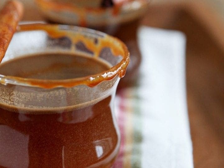Two glasses of Spiked Caramel Apple Cider are served on a wooden platter, rimmed with caramel and garnished with a cinnamon stick