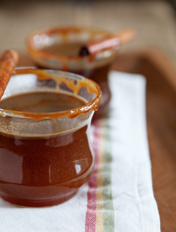 Two glasses of Spiked Caramel Apple Cider are served on a wooden platter, rimmed with caramel and garnished with a cinnamon stick